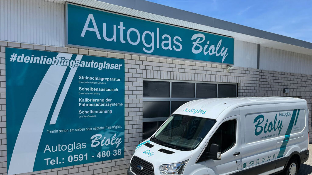 Ein weißer Autoglas-Wagen vor einem Laden mit großen Schildern und Schriftzug "#deinlieblingsautoglaser".